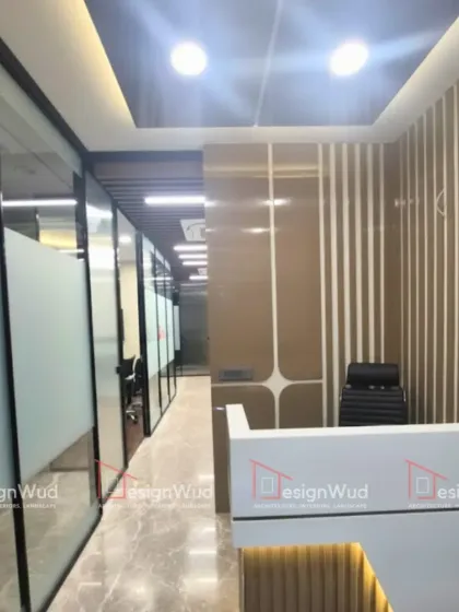 The reception area of a corporate office in Noida, designed to make a strong first impression. It features a sleek white reception desk with accent lighting, set against a backdrop of vertically grooved and glossy wall panels. The glass partitions provide a glimpse into the workspace beyond.