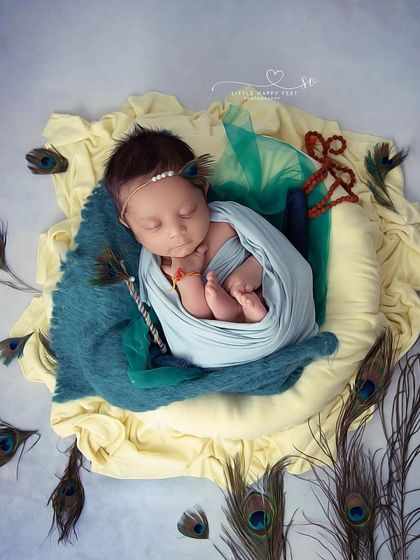Another beautiful Krishna-themed portrait, with the baby wrapped in blue and yellow, surrounded by a cascade of peacock feathers. This top-down angle creates a lovely, encompassing feel.