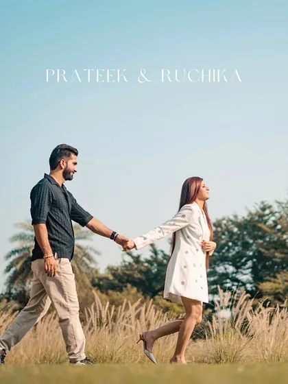 A stylish shot of the couple walking through a field of tall grass under a clear blue sky. This pre-wedding photo has a chic, editorial feel.