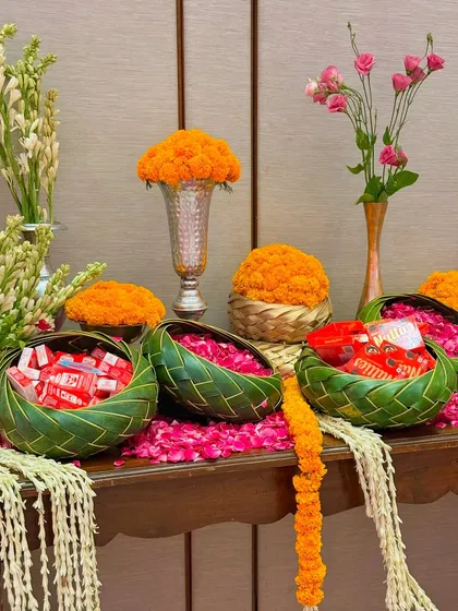 A festive table arrangement featuring bowls of marigolds, rose petals, and traditional Indian sweets. The use of natural leaf bowls and tuberose garlands adds an organic and authentic touch to the decor.