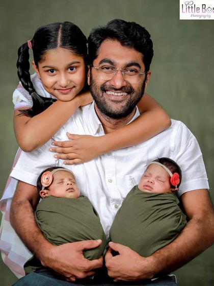 A proud father with his three children. He holds his newborn twins while his older daughter gives him a loving hug from behind.