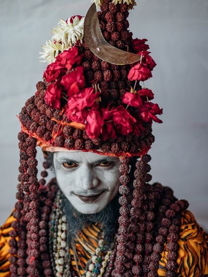 A striking portrait of a Naga sadhu at the Kumbh Mela, his face painted and adorned with a crown of Rudraksha beads and roses. His expression is both fierce and captivating.