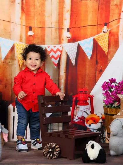 A happy one-year-old boy in a cozy rustic cafe setting, complete with a tiny chair, wooden fence, and string lights.