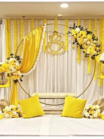 A bright and sunny Haldi decor setup featuring a circular golden frame backdrop. The look is completed with yellow drapes, marigold garlands, and a comfortable bench for the ceremony.