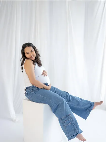 A relaxed and happy portrait. Sitting comfortably in jeans and a tank top, this mom-to-be radiates a calm and confident energy against a simple white backdrop.