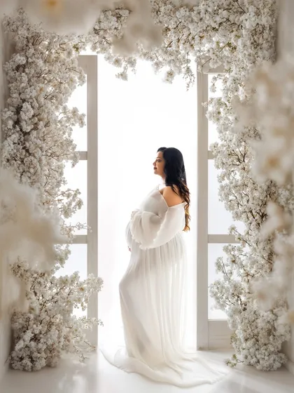 This is the single most important photo to capture your pregnancy glow. Here, a stunning mommy-to-be stands in a flowing white gown, framed by a beautiful arch of white blossoms, creating a timeless and ethereal maternity portrait.
