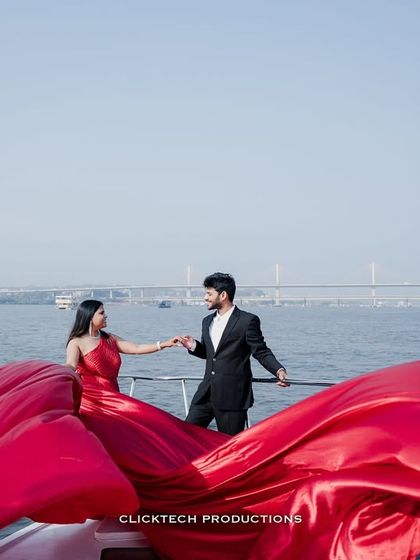 A wide shot of a couple on a yacht in Goa, the massive red trail of her gown spread across the deck, with the sea and bridge in the background.