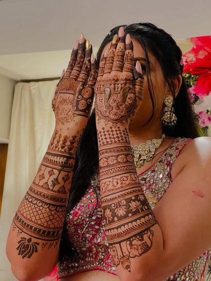 A classic bridal pose by Raksha, her hands framing her face, highlighting the detailed henna work.
