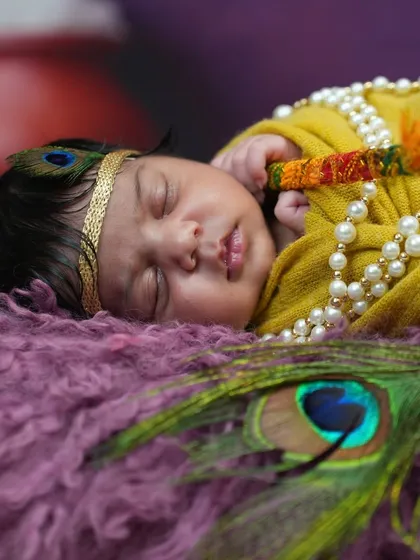A sleeping newborn becomes the divine Kanha, adorned with a simple peacock feather and pearl necklace, showcasing a softer take on the Krishna theme.