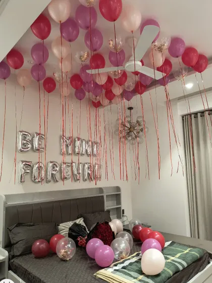 A promise of love with 'BE MINE FOREVER' foil balloons. The ceiling is covered with pink, magenta, and confetti balloons, creating a festive and deeply romantic atmosphere in the bedroom.
