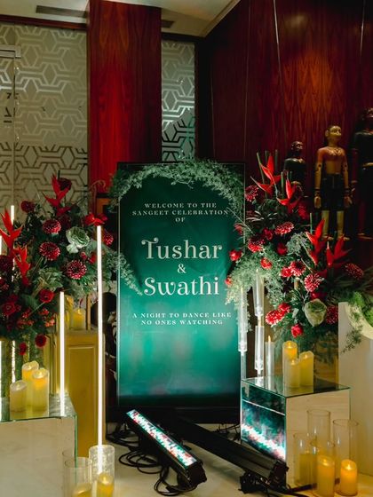 A chic welcome sign for a Sangeet celebration, set against an emerald green background. The sign is surrounded by modern lighting and bold red floral arrangements, perfectly matching the event's glam theme.
