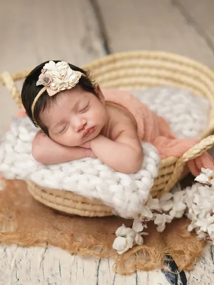 A newborn girl rests her head on her hands in a soft basket. The combination of textures, from the knit blanket to the wooden floor, adds depth to this infant portrait.