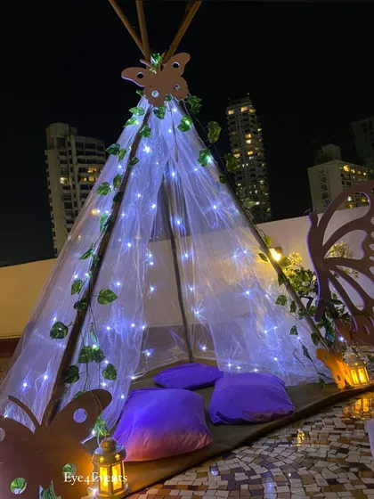 A wider shot of the butterfly-themed teepee against the city backdrop at night. The cool-toned fairy lights and purple cushions tie back to the main lavender theme of the party, creating a cohesive look.