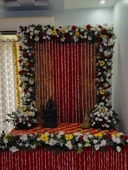 A simple yet elegant floral backdrop for a home ceremony or puja, created with a frame of fresh flowers and a red and white striped cloth.
