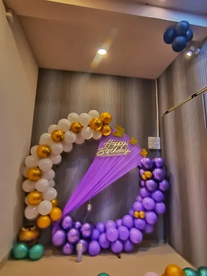 A corner birthday setup with a circular balloon arch in purple, white, and gold. A purple drape and butterfly accents complete this charming decoration.