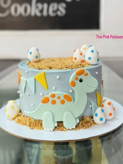 A playful dinosaur cake with a brontosaurus figure and speckled dinosaur eggs. The edible sand and simple banner complete this prehistoric-themed design.