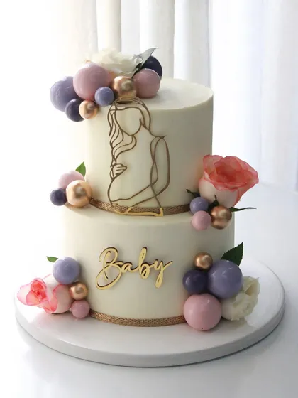 An elegant two-tiered baby shower cake with a "mom-to-be" silhouette, fresh flowers, and a cascade of colorful chocolate spheres.