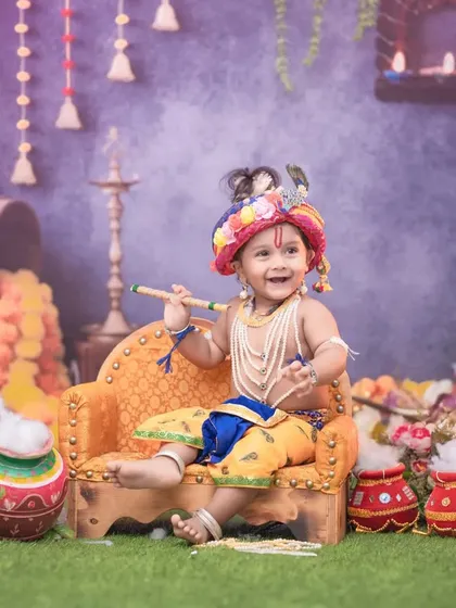 A whimsical Krishna-themed setup with a baby boy playing a flute on a miniature couch. The background is filled with festive elements like marigold garlands, earthen pots, and soft lighting.