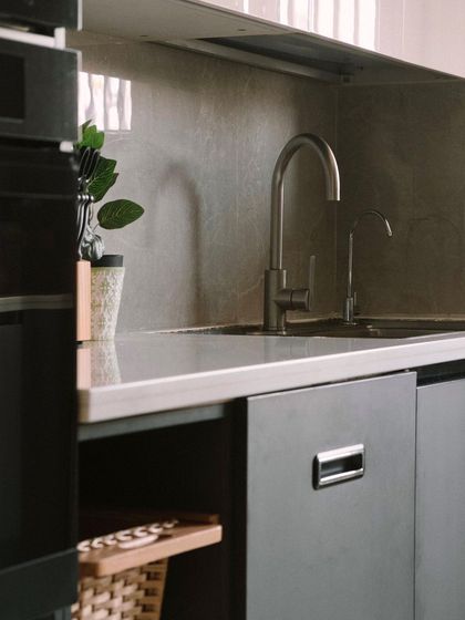 A detail of a modern kitchen, focusing on the sleek brushed metal faucet and the subtle texture of the stone backsplash, reflecting a minimalist and clean aesthetic.