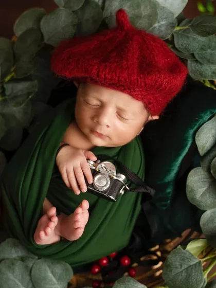A tiny artist in the making! This creative newborn portrait features a baby in a red beret, holding a miniature camera, nestled amongst lush green leaves.