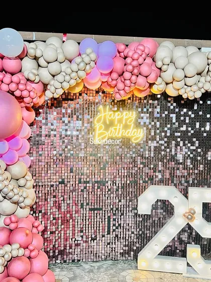We designed this glamorous photo backdrop for a 25th birthday celebration. The setup combines a sparkling rose gold shimmer wall with a custom 'Happy Birthday' neon sign and large, light-up marquee numbers to create a perfect spot for party pictures. The scene is framed with an organic balloon arch in shades of pink and beige.