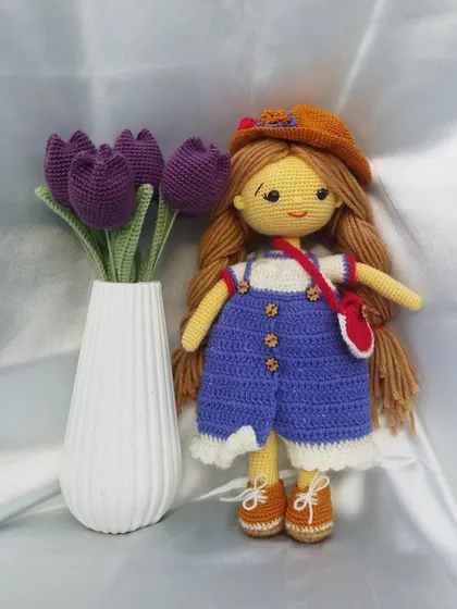 A custom-made crochet doll with long brown hair, wearing blue overalls and a hat. She stands next to a vase of my purple crochet tulips, giving a sense of the doll's size and the detail I put into each character.