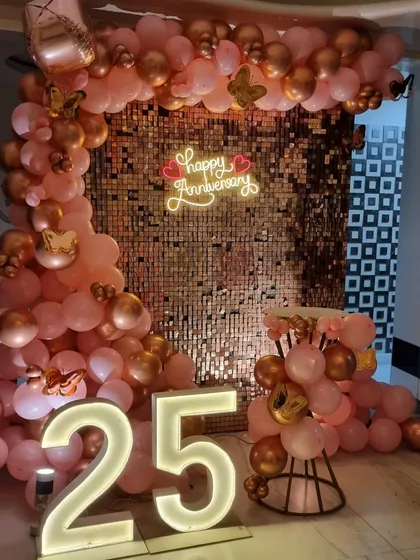 A glamorous 25th-anniversary decoration featuring a rose gold sequin wall. The setup is framed by a pink and gold balloon arch and highlighted by large illuminated "25" numbers.