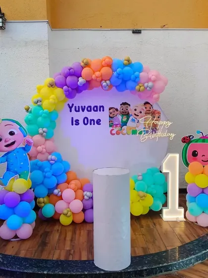 A vibrant Cocomelon first birthday decoration for "Yuvaan". This outdoor setup features a circular backdrop, character pillars, a neon sign, and a light-up number "1".