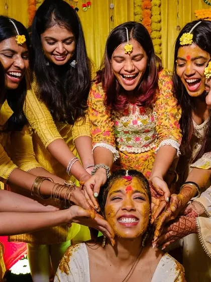 The bride is all smiles, surrounded by her laughing friends as they playfully apply haldi to her face.