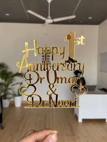 The "Happy 1st Anniversary Dr. Umar & Dr. Noori" topper held up to show its full design and reflective surface.