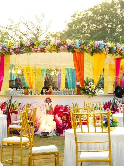 A lively outdoor bar setup for a mehandi or sangeet. The counter is decorated with bold floral graphics, and the overhead structure is adorned with colorful flowers and drapes, adding to the festive atmosphere.