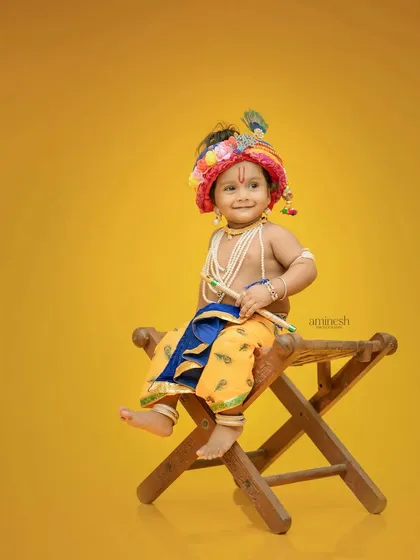 A simple and sweet portrait of a baby dressed as Krishna, seated on a wooden stool. The solid yellow background keeps the focus entirely on the child's adorable outfit and expression.