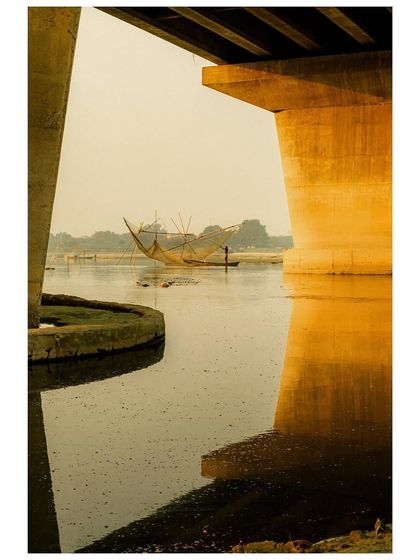 A fisherman in his boat casts a large, traditional Chinese fishing net on the Yamuna River, framed by the concrete pillars of a bridge. The shot highlights the contrast between old and new methods of life.