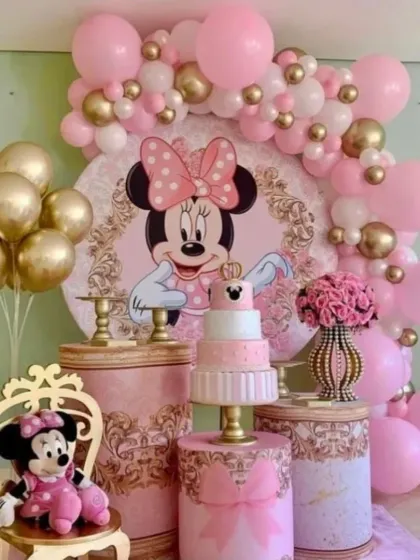 A classic and elegant Minnie Mouse first birthday. This setup uses a pink and gold color palette, with themed plinths for the cake, a character backdrop, and a mini throne for the birthday girl.