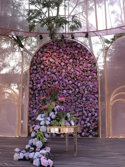 A stunning floral wall made of purple and blue hydrangeas serves as a backdrop. The cascading flowers on the table create a sense of organic, overflowing beauty.