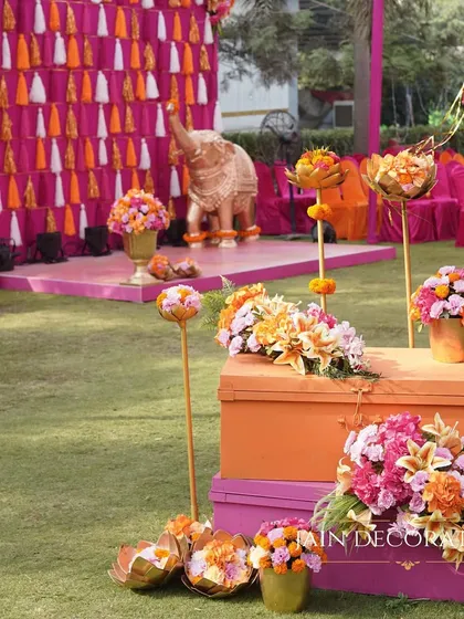 A beautiful blend of vibrant colors for a daytime function. The lawn is dotted with floral arrangements in pink and orange, with a traditional elephant statue and a tassel wall adding to the festive decor.