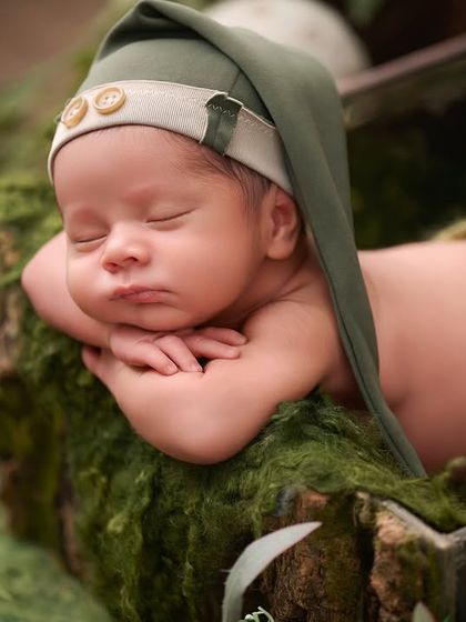 A baby wearing a green pixie hat sleeps in a moss-lined wooden box, creating a magical woodland creature portrait.