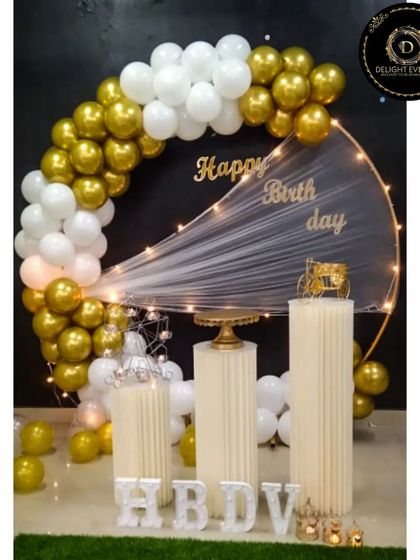 An elegant and sophisticated setup for a large space. This design uses a gold and white balloon arch with a sheer, lit fabric drape, paired with classic pedestals and light-up initials for a clean and luxurious feel.