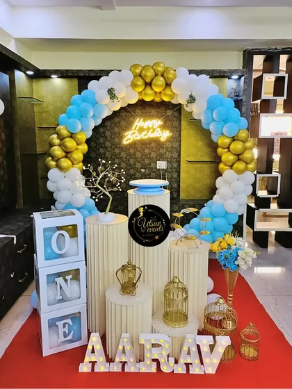 A first birthday celebration with a blue, white, and gold balloon ring. The setup includes 'ONE' blocks and a crown prop for the little prince.
