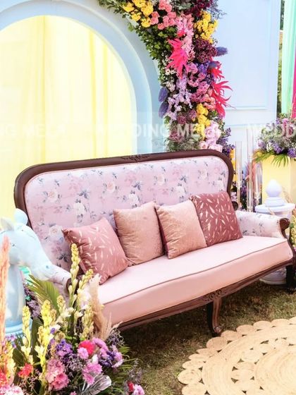 A beautiful and whimsical mehandi seating area. The vintage-style floral sofa is framed by a stunning arch of vibrant, multi-colored flowers, creating a perfect photo backdrop.