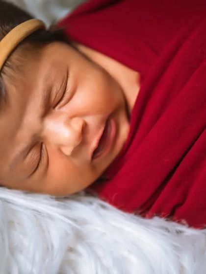 A close-up of a newborn with a sweet expression, wrapped in a red swaddle on a white fur blanket. The simple red headband adds a touch of color.