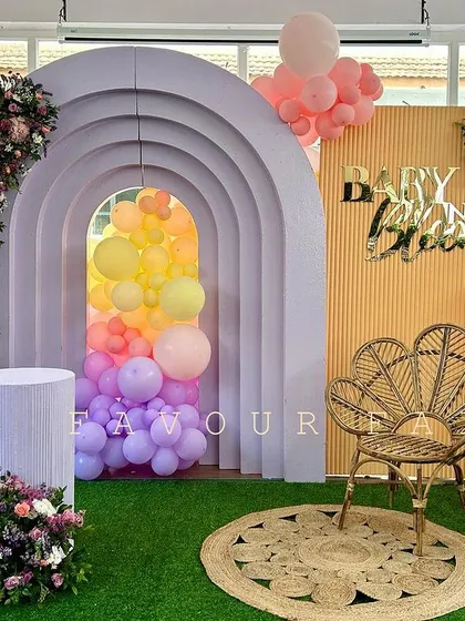 A 'Baby in Bloom' baby shower with a rainbow of pastel colors. The setup features a unique arched backdrop with a balloon-filled tunnel effect, a wicker peacock chair, and beautiful floral arrangements.