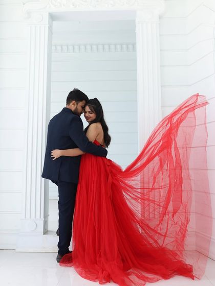A romantic pre-wedding embrace in a studio with an all-white background. The flowing red trail of the gown provides a stunning pop of color.