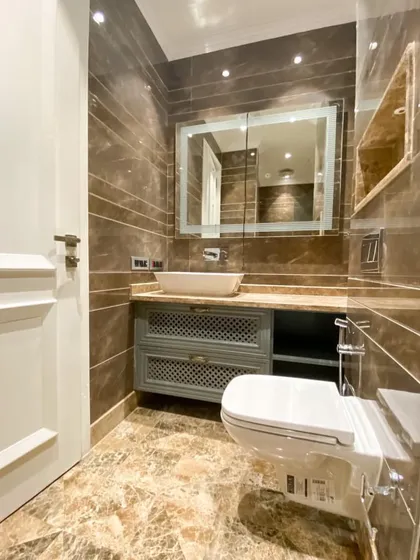 A beautifully designed bathroom featuring a custom grey vanity with latticework doors, a modern vessel sink, and an illuminated mirror, all set against rich brown marble.
