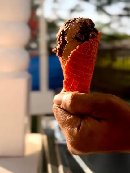 A hand holds a scoop of rich chocolate gelato in a crisp waffle cone against a sunny backdrop. It’s a perfect picture of a delightful, refreshing treat enjoyed at an outdoor event.