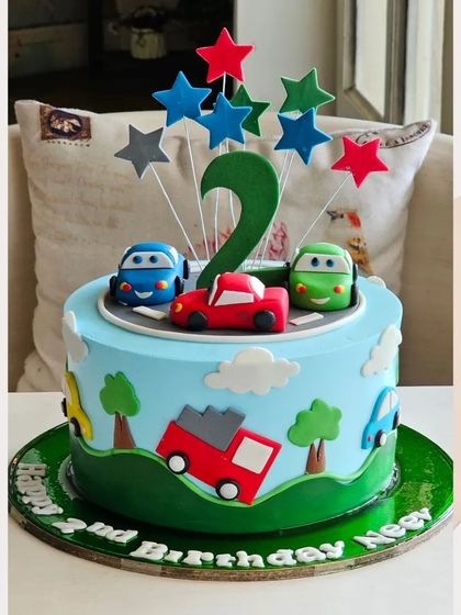 A colorful cake for a 2nd birthday, featuring a road with cute cartoon cars and trucks driving through a green landscape.