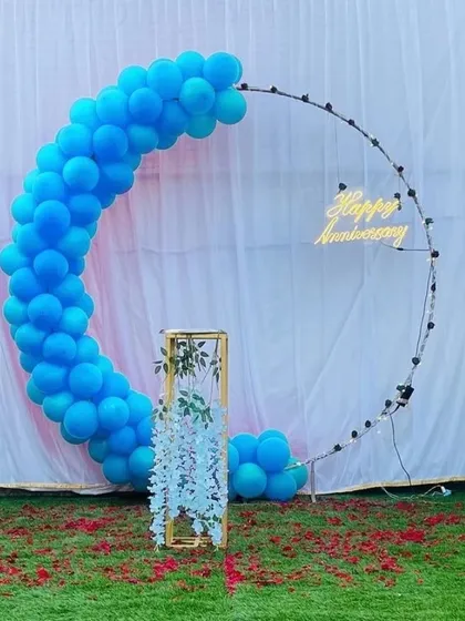 A minimalist and modern anniversary backdrop. A simple blue balloon crescent on a circular frame is paired with a neon sign and a floral arrangement in a gold stand.