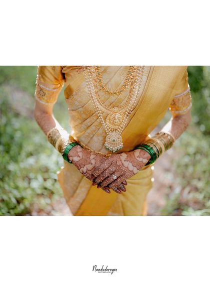 An artistic shot focusing on the bride's hands, showcasing her intricate henna, green bangles, and the stunning gold necklace against her silk saree.