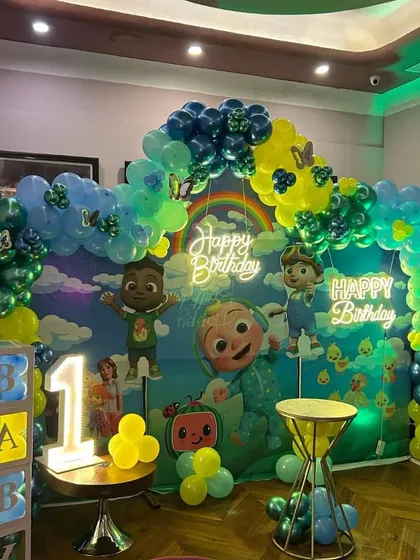 A Cocomelon first birthday setup with a large themed backdrop, a '1' marquee light, and 'BABY' balloon boxes.
