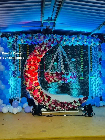 A magical fairy-themed naming ceremony stage. The design includes a floral crescent moon with a hanging cradle, set against a green backdrop with blue balloon pillars and fairy cutouts.